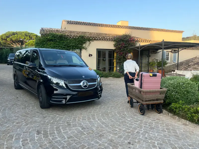 Mercedes Classe V noir Cannes Driver stationné devant une villa provençale au coucher du soleil, chargement de valises roses sur chariot en bois par une voyageuse